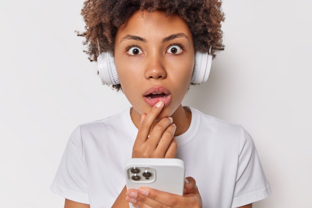 Portrait of startled young woman gazes surprised has bugged eyes listens music via stereo headphones holds mobile phone dressed casually isolated over white background Human reactions concept – VidAU.ai A woman surprised after realizing she can download YouTube audio