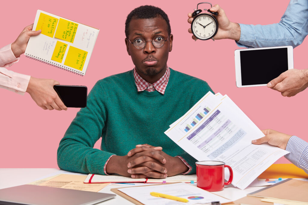 Sad young African American man dressed in green sweater sits at desktop hands with papers alarm clock touchpad notepad with stickes studies documentation isolated over pink background