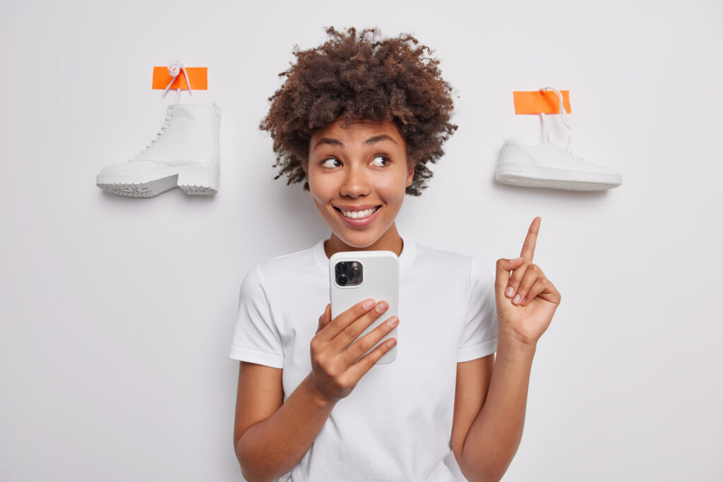 Waist up shot of glad brunette woman points index finger above on plastered shoes smiles toothily recommends something to wear uses mobile phone surfs online store isolated over white studio wall shop sneaker deals women's