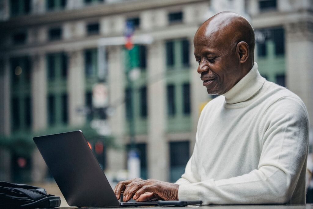 Side view of self employed concentrated mature bald black man in stylish turtleneck sitting in street cafe and typing on netbook during work